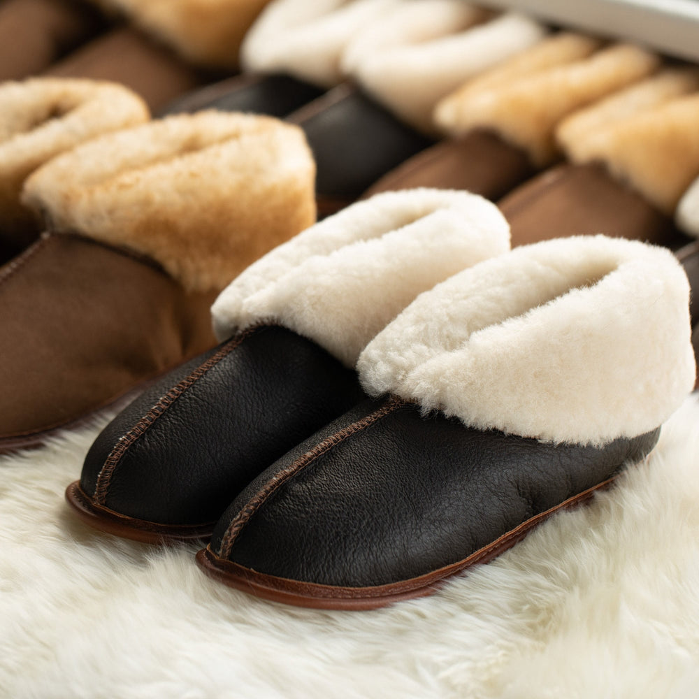 Close up shot of two rows of Ithaca Sheepskin slippers displayed on an ivory sheepskin rug. Front and center is a pair of slippers with dark brown leather uppers, creamy shearling fur visible on folded down ankle cuffs