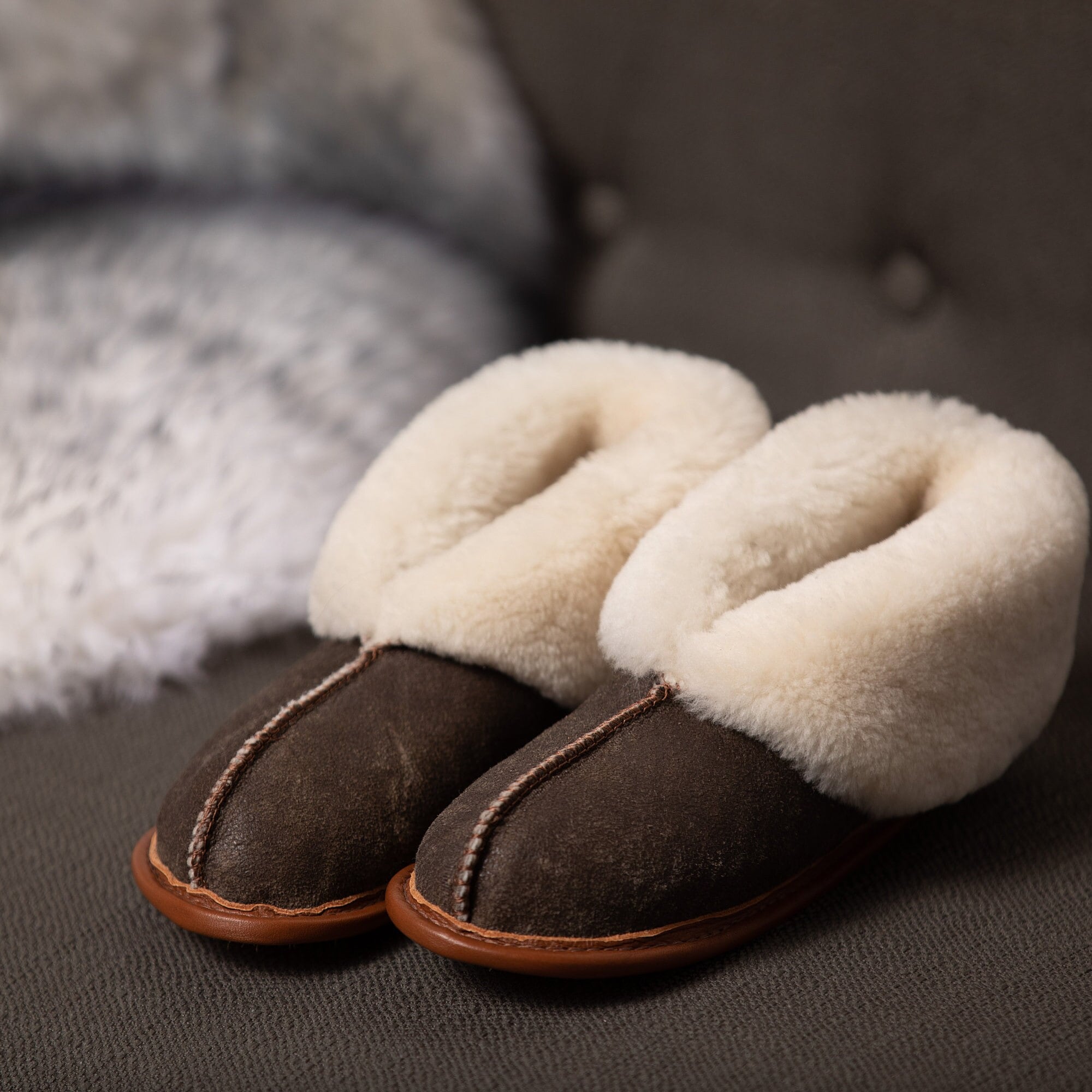 A brown pair of Ithaca Sheepskin slippers with the cuffs down to expose the cream colored fur lining, on a brown couch with a white and gray fleece in the background
