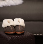 A pair of brown Ithaca Sheepskin slippers with the cuffs folded down to expose the cream colored shearling fur lining. The slippers are on an ottoman, in front of a sofa with a gray and white fleece on it.