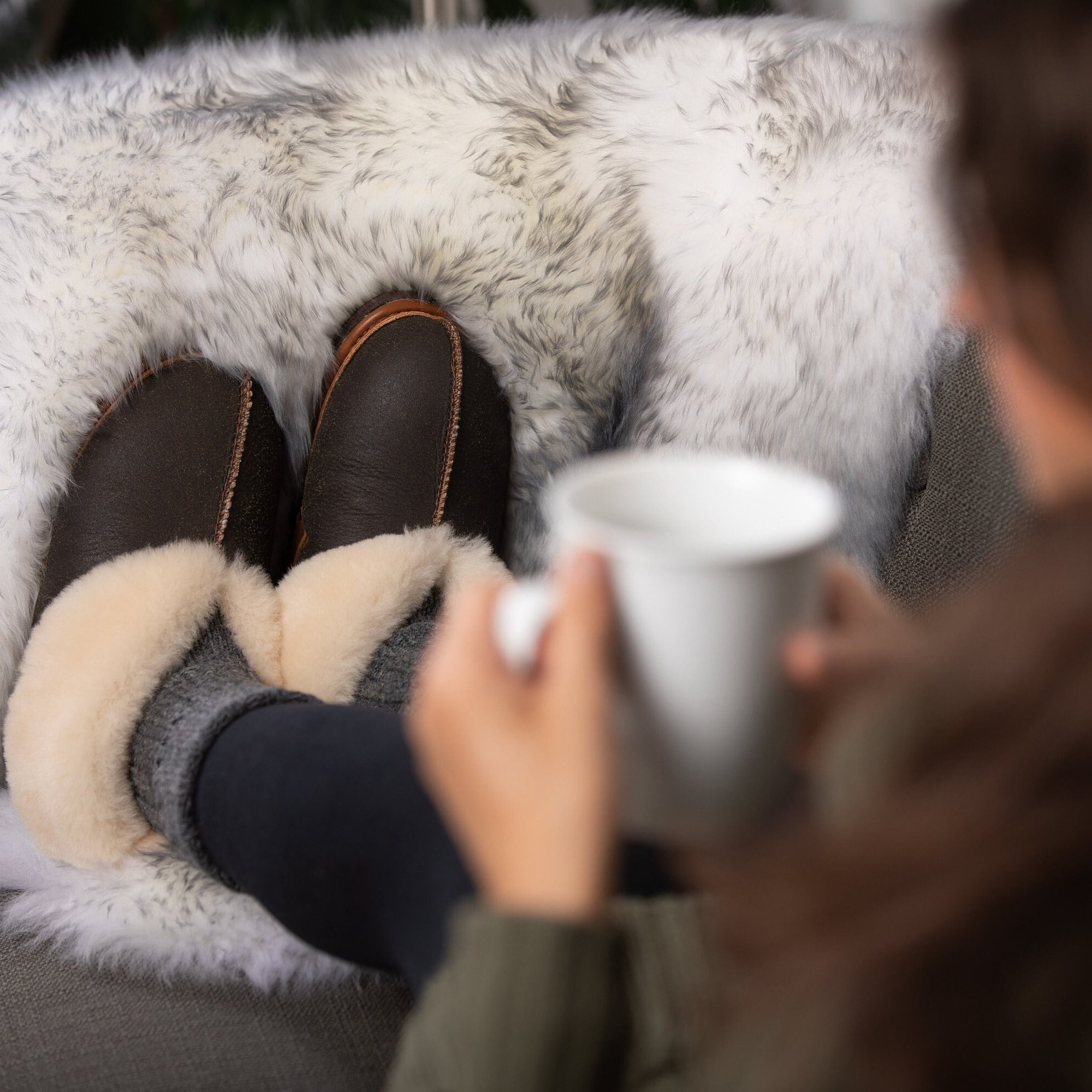 A person sits on a couch covered with fleeces, wearing a pair of brown Ithaca Sheepskin slippers and enjoying a coffee.