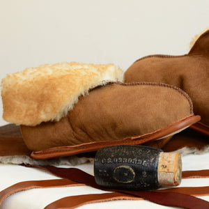A beige pair of Ithaca Sheepskin slippers, with brown tipped wool exposed on folded cuff. The slipper in the foreground is propped on a mallet, with strips of leather binding laying on the surface beneath.